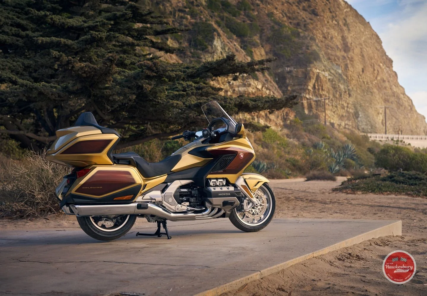Gold and black Honda Gold Wing touring motorcycle parked on a concrete pad in front of a desert landscape with hills, shrubs, and a blue sky.