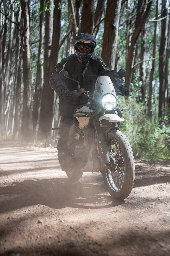 Person riding a black and white motorcycle on a dirt trail through a forest, wearing a black helmet and gear.