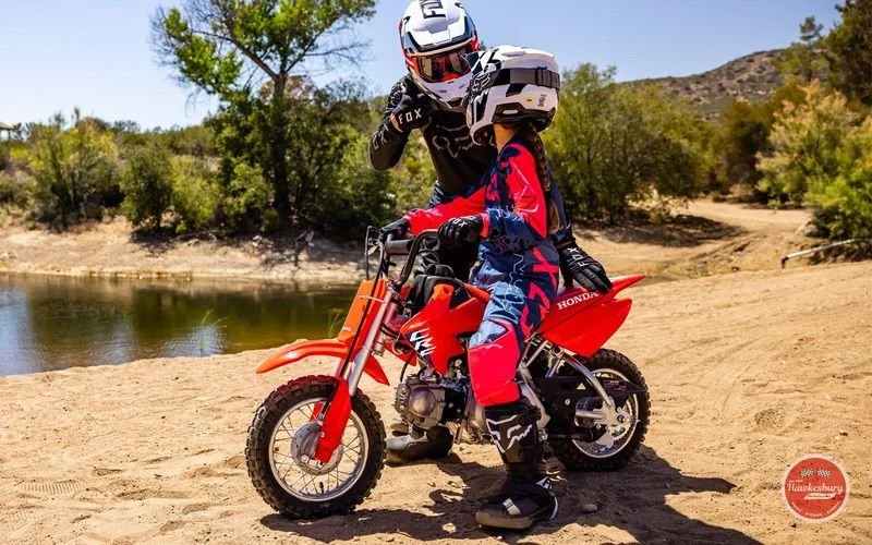 A young child dressed in motocross gear sitting on a red dirt bike with an adult helping them hold the handlebars on a dirt trail next to a river, with trees and hills in the background.