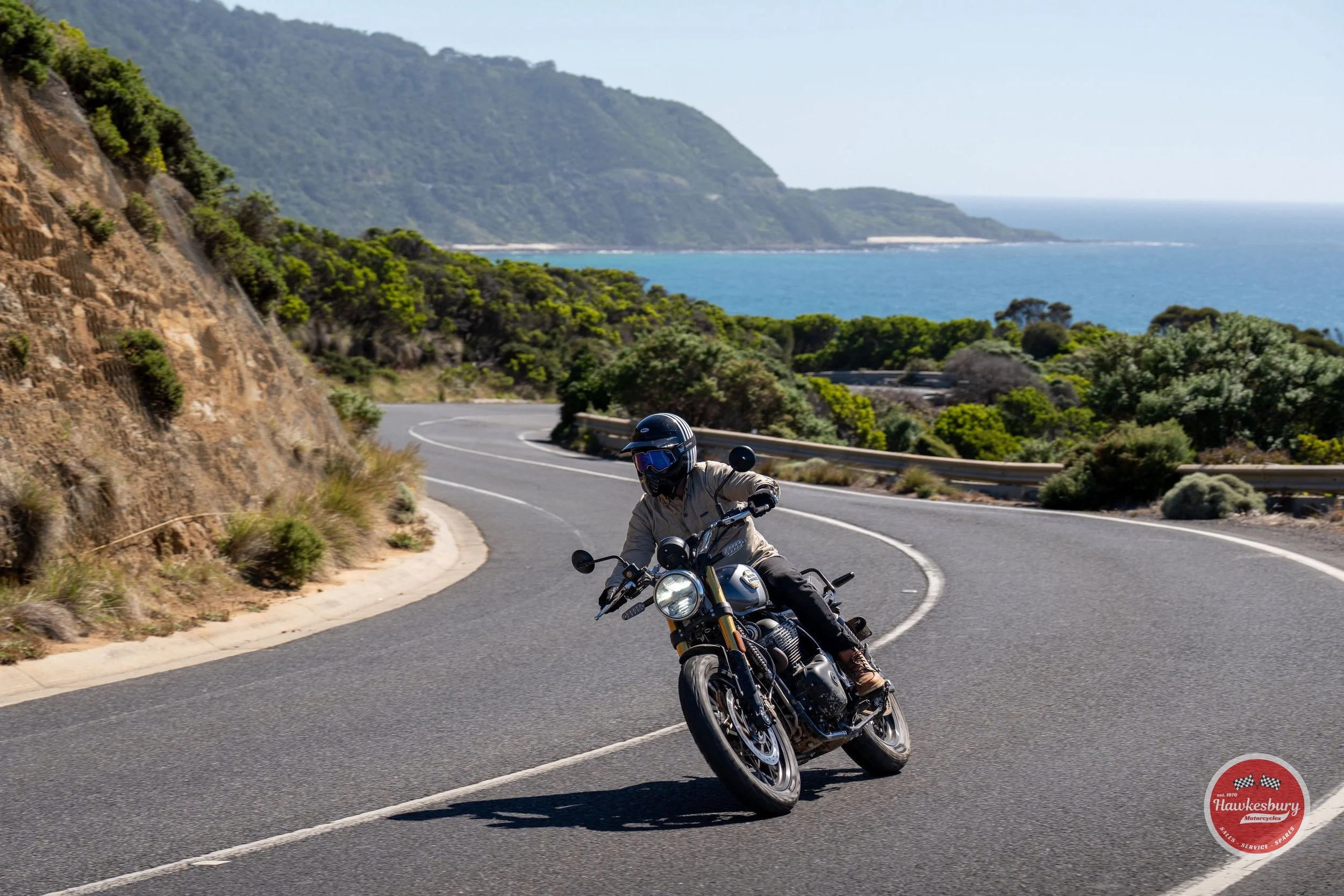 Motorcycle rider wearing a helmet and beige jacket leaning into a curve on a scenic coastal mountain road with the ocean in the background.