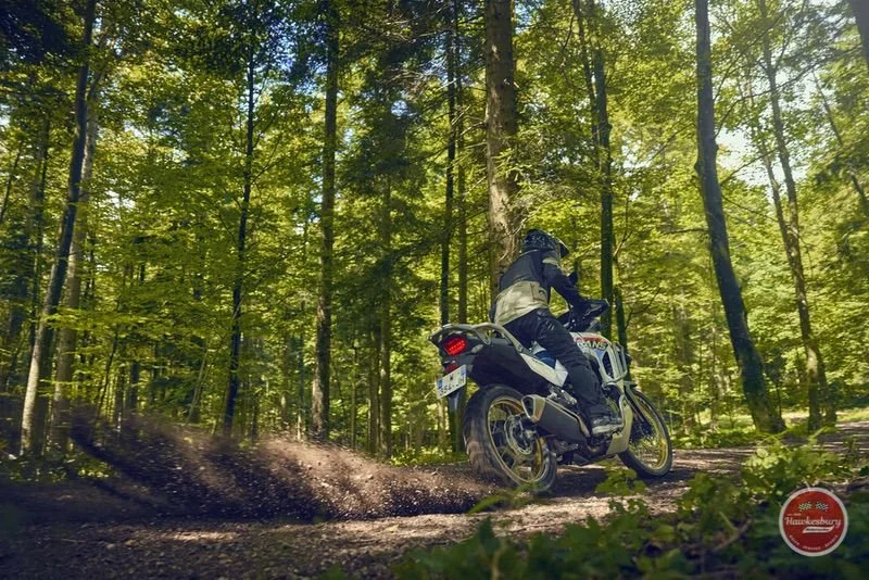 Motorcycle rider wearing black gear riding through a green forest on a dirt trail, kicking up dirt behind the bike.