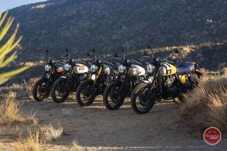 Five vintage-style motorcycles lined up on a dirt trail in a mountainous area with dry grass and bushes.