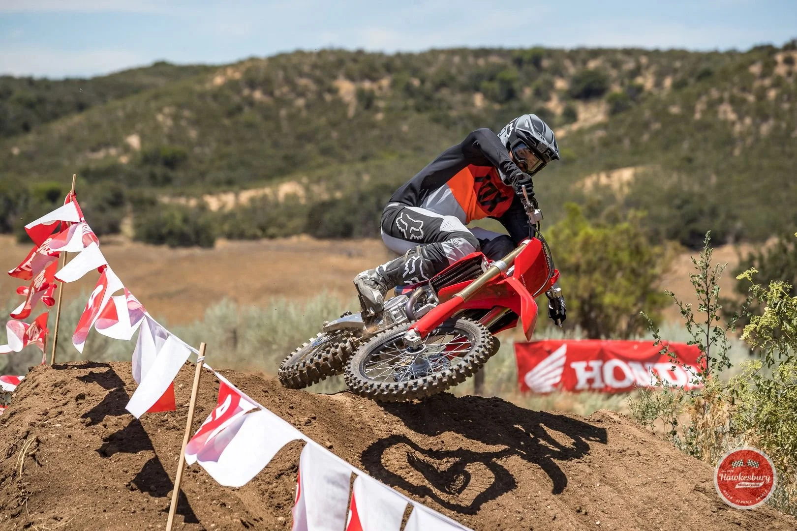 A motocross rider in black, gray, and red gear maneuvers on a red dirt bike over a dirt mound during a race, surrounded by red and white flags and Honda banners, with a scenic landscape of hills and sparse vegetation in the background.