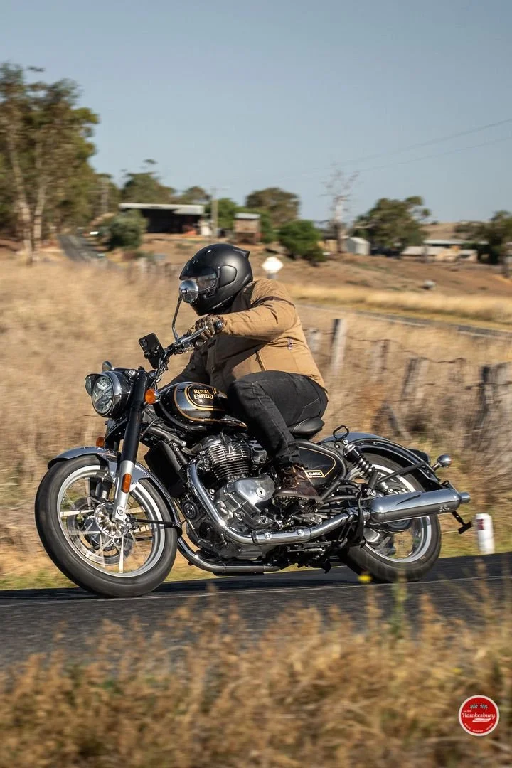 A person riding a black Royal Enfield motorcycle on a rural road, wearing a brown jacket and black helmet, with a rural landscape in the background.