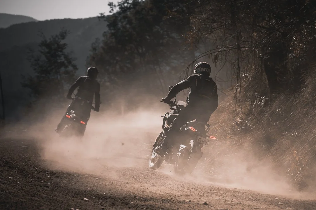 Two motorcyclists riding on a dirt trail in a wooded area during dusk or dawn, kicking up dust as they navigate the path.