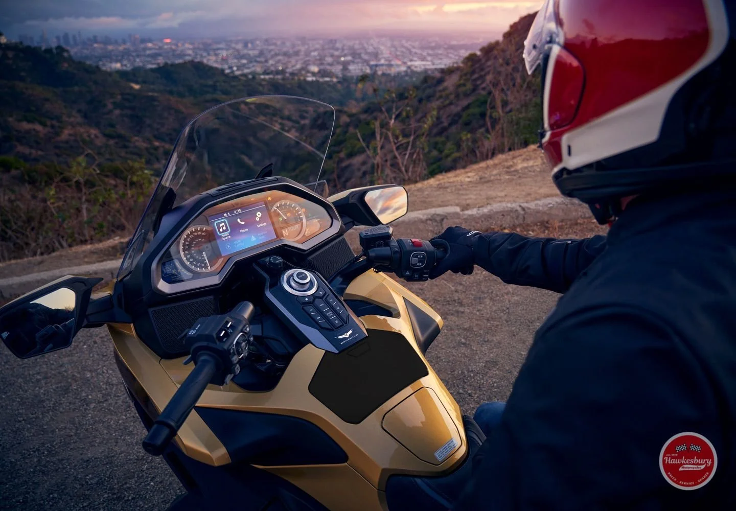 A motorcyclist wearing a red helmet and black jacket, riding a gold and black motorcycle on a mountain road during sunset, with a cityscape in the background.