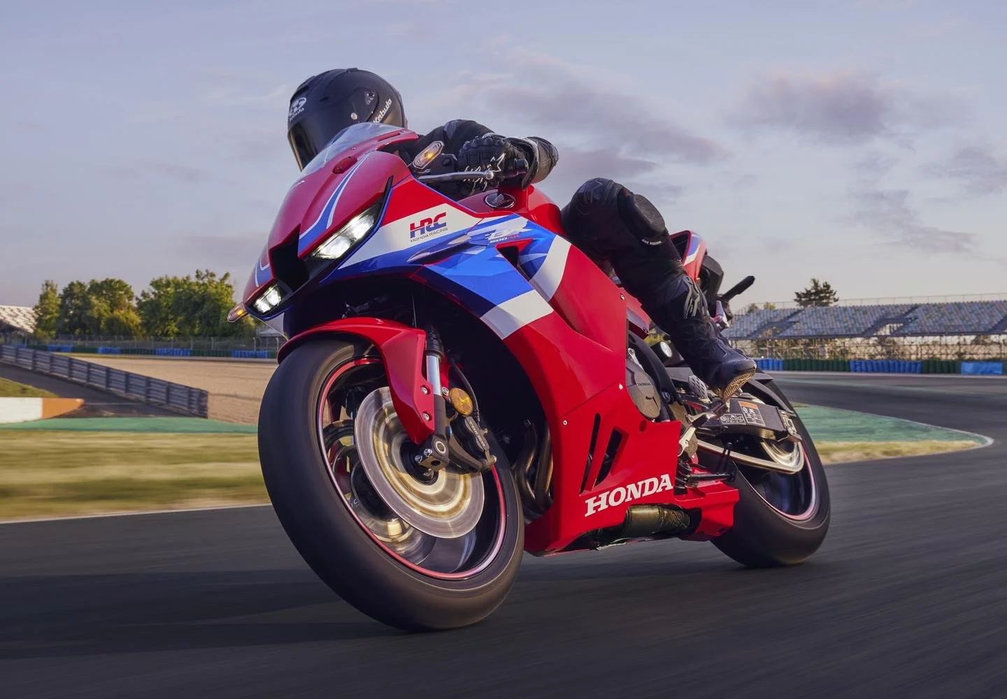 A red, blue, and white Honda racing motorcycle in a turn on a race track, with a rider wearing black protective gear and a helmet leaning into the curve during daytime.