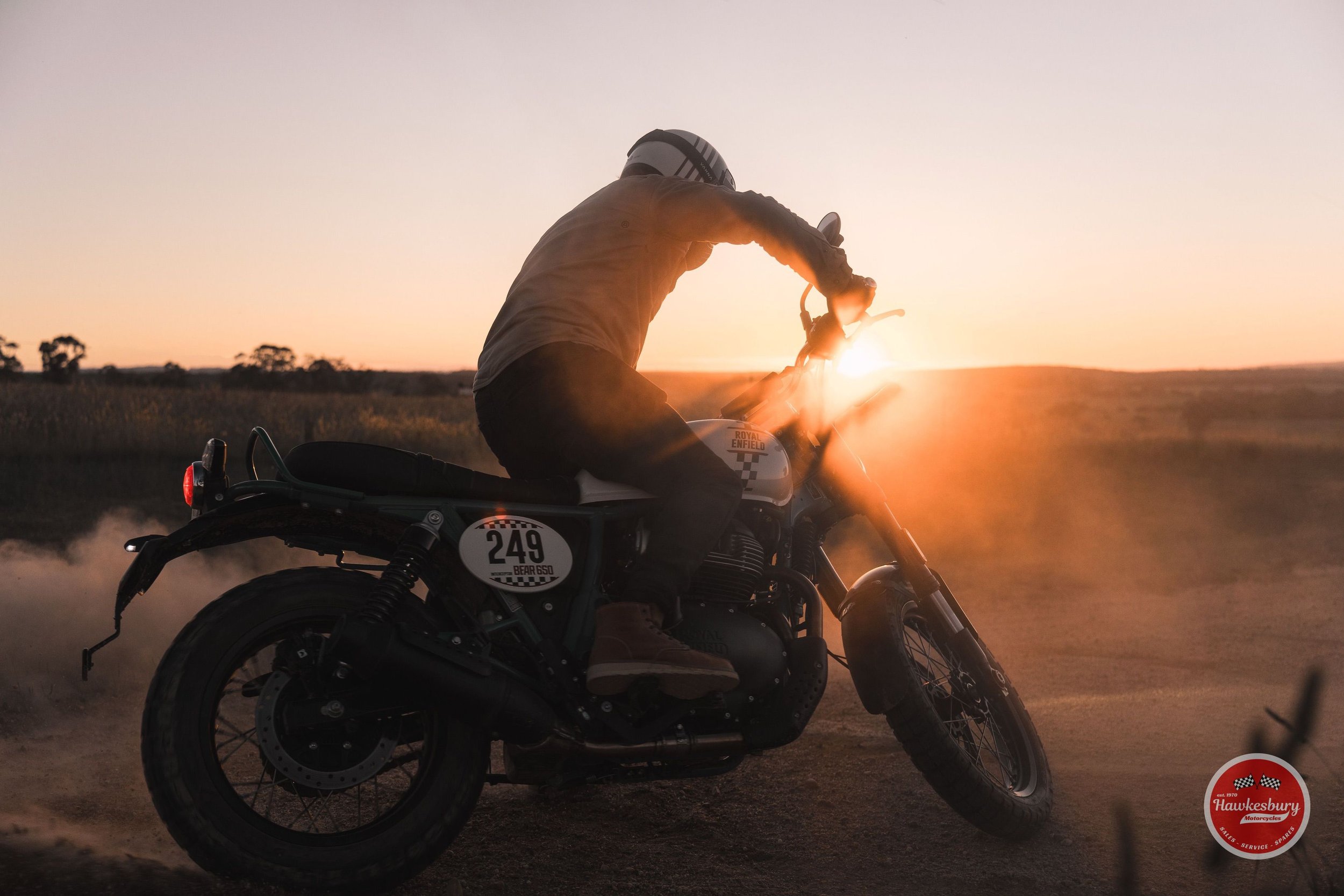 A person riding a motorcycle on a dirt road during sunset, with a dust cloud behind them and the logo 'Hawkesbury Motorcycles' in the bottom right corner.