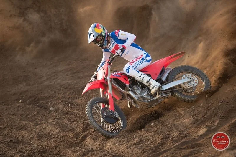 A motocross rider wearing a white, blue, and red suit and helmet riding a red dirt bike on a dirt track.