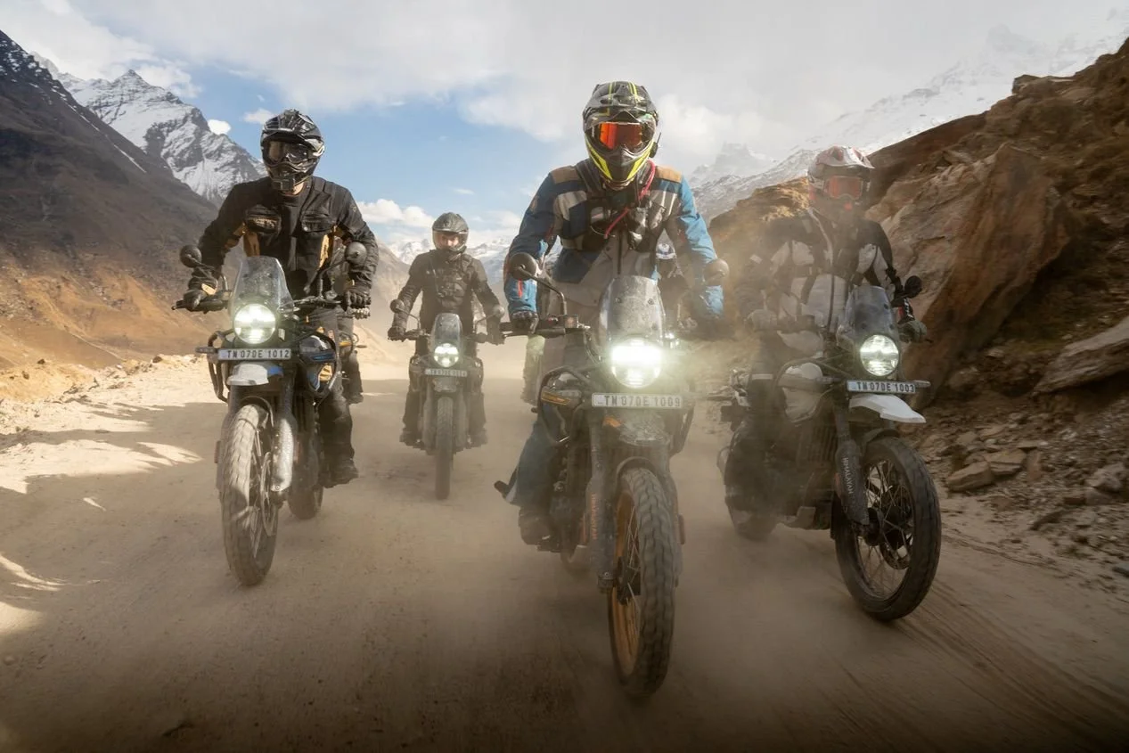 Group of five motorcycle riders traveling on a dusty mountain road with snow-capped peaks in the background.