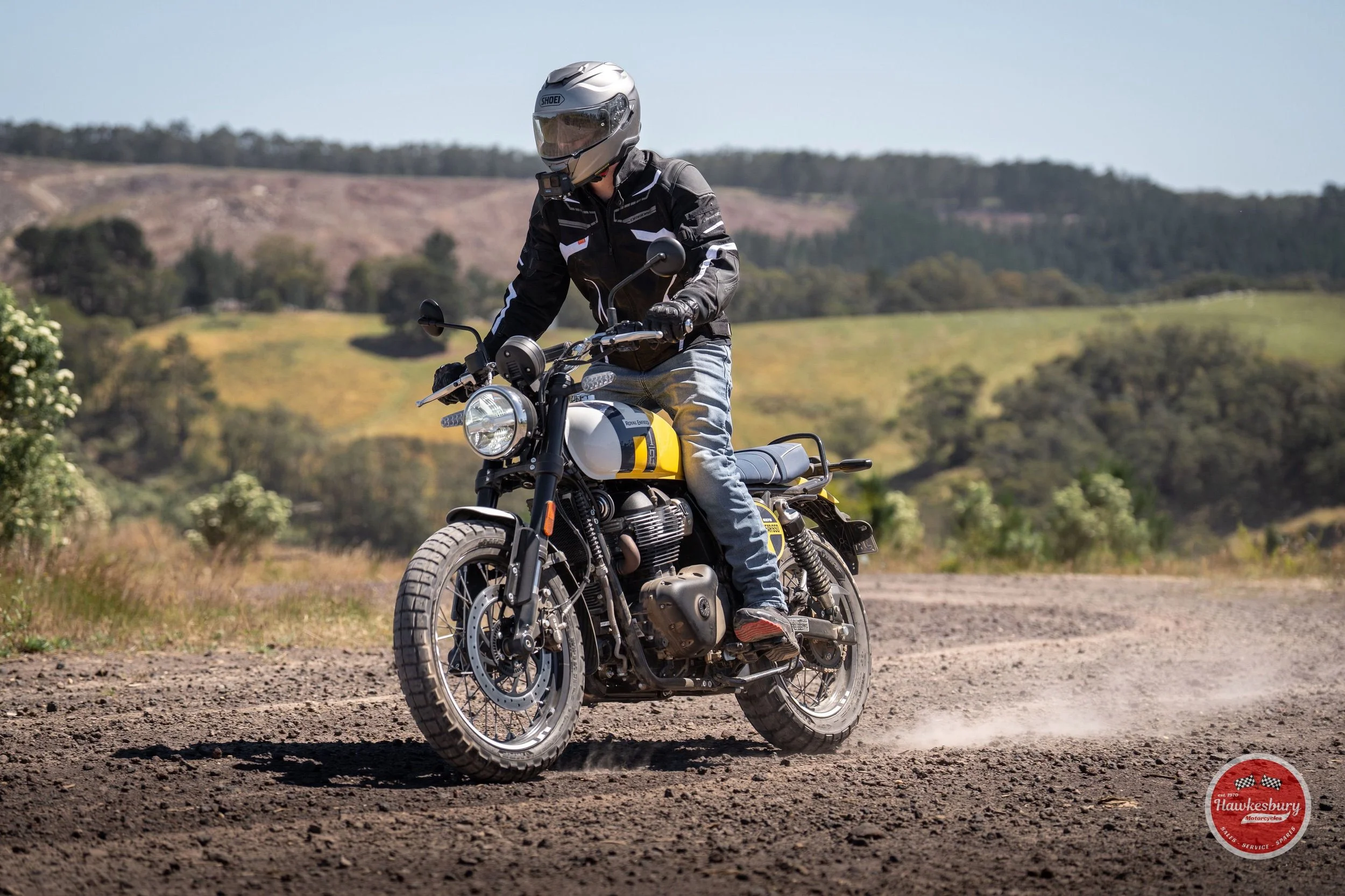 A person riding a motorcycle on a dirt road in a rural area with green hills and trees in the background.