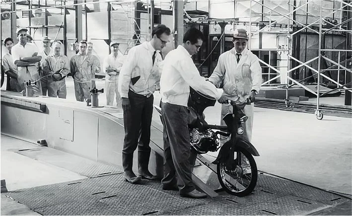 Technicians and engineers in a factory setting inspecting a motorcycle on a testing device, with scaffolding and equipment in the background.