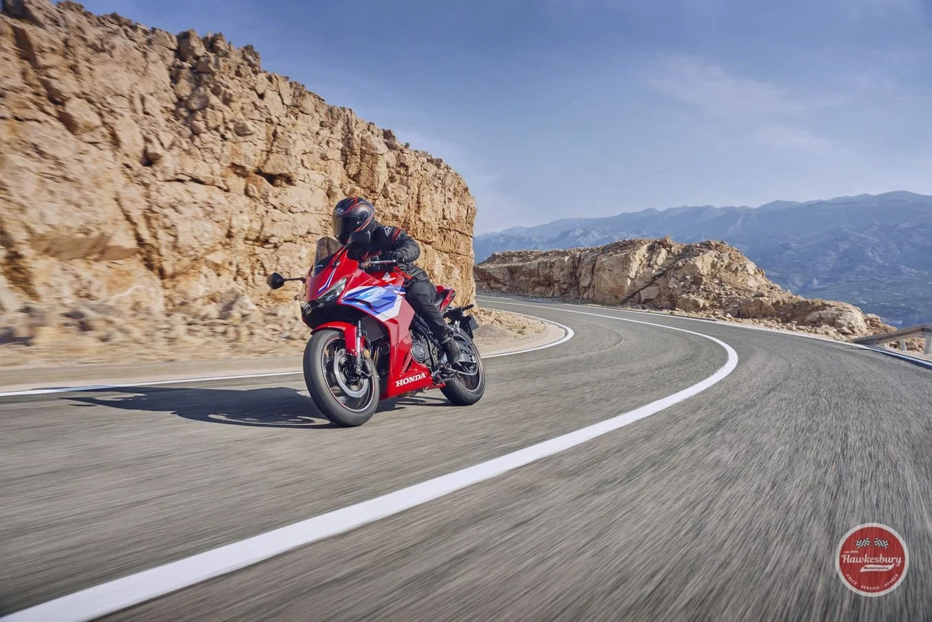 Motorcycle rider on a winding mountain road with rocky cliffs and distant mountains in the background.
