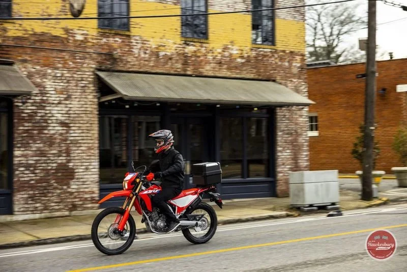 A person wearing a helmet riding a red and white motorcycle on a city street, with brick buildings and a tree in the background.