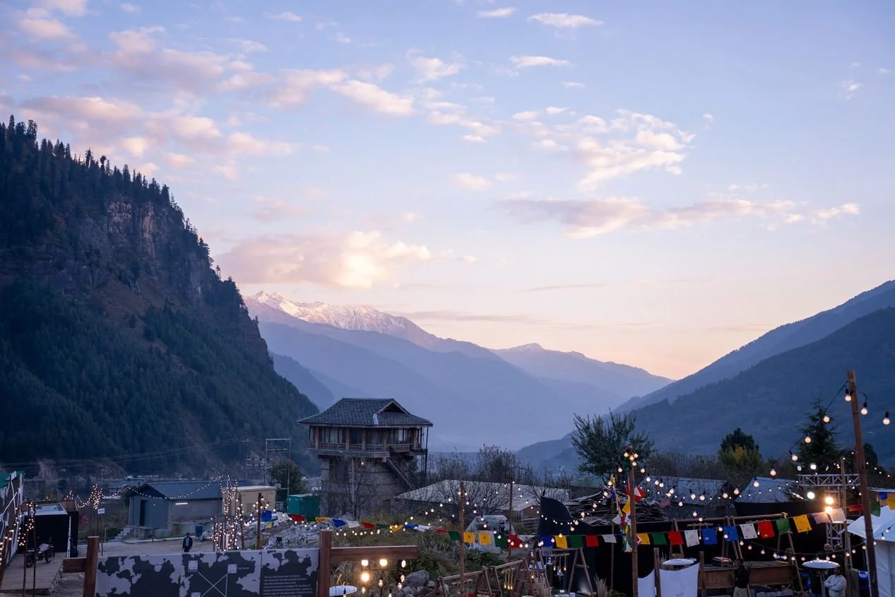Alpine mountain village at dusk with string lights, tents, and a wooden house, surrounded by tall mountains and a cloudy sky.