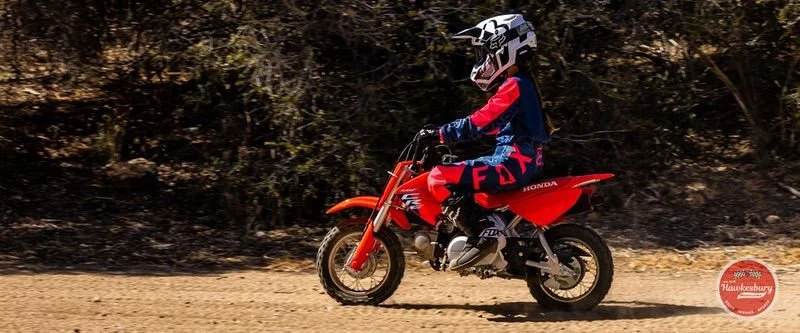 A person riding a red Honda dirt bike on a dirt trail with trees in the background, wearing a helmet and motocross gear.