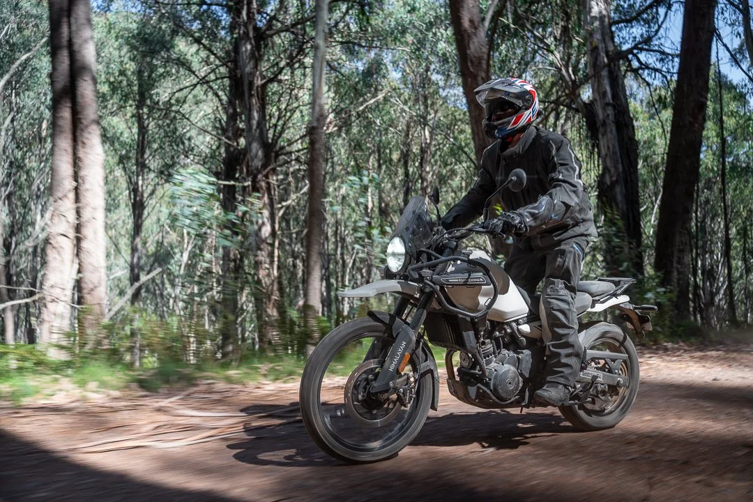 A person riding a motorcycle through a wooded trail, wearing a black helmet with a Union Jack design and a black riding jacket.