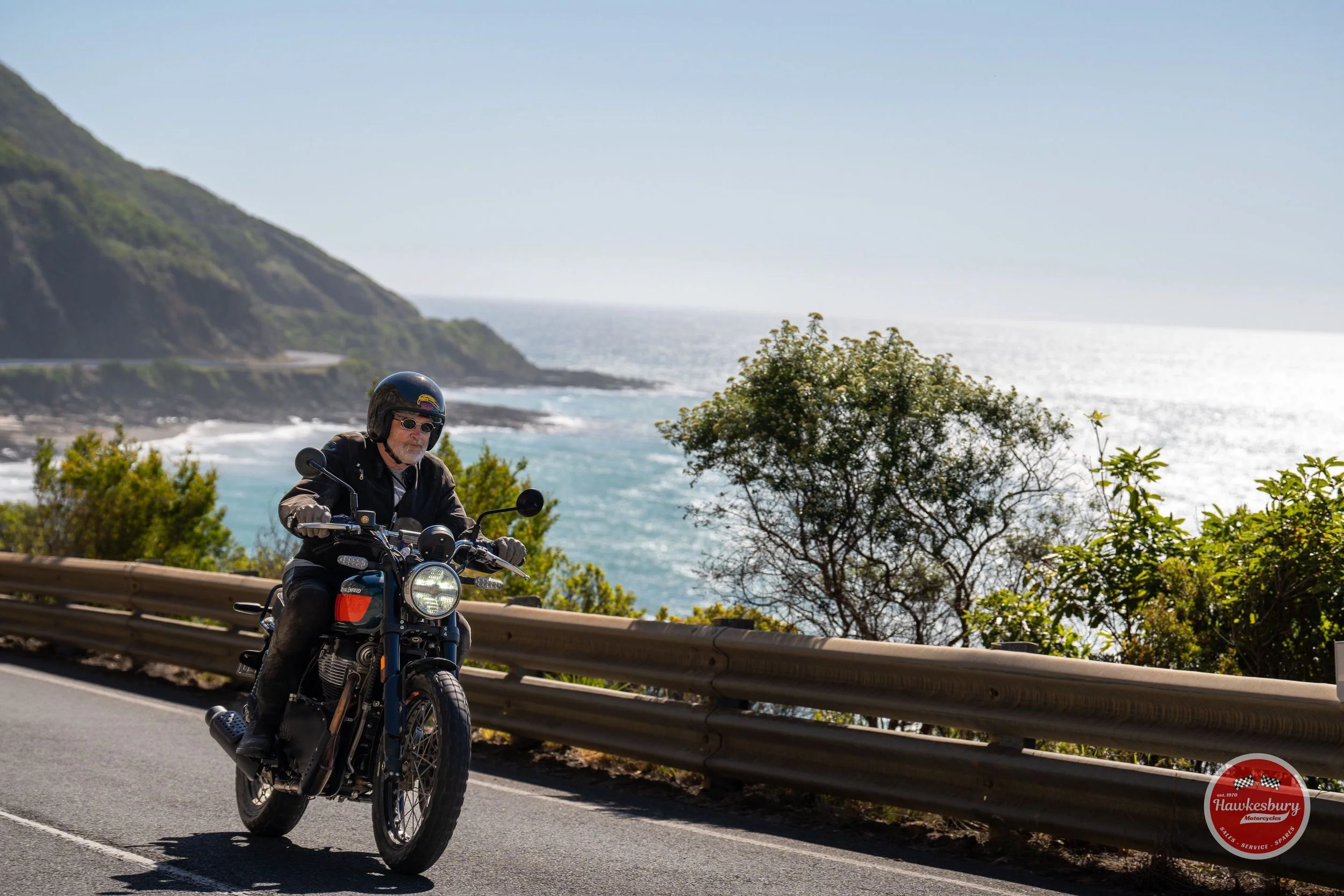 A man riding a motorcycle along a coastal road with the ocean, hills, and trees in the background during the day.