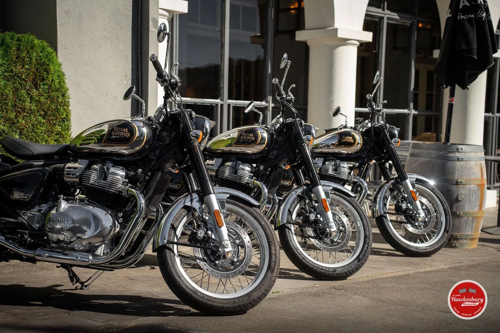 Three black and chrome Royal Enfield motorcycles parked outside a building on a sidewalk, with a bush and a barrel in the background.