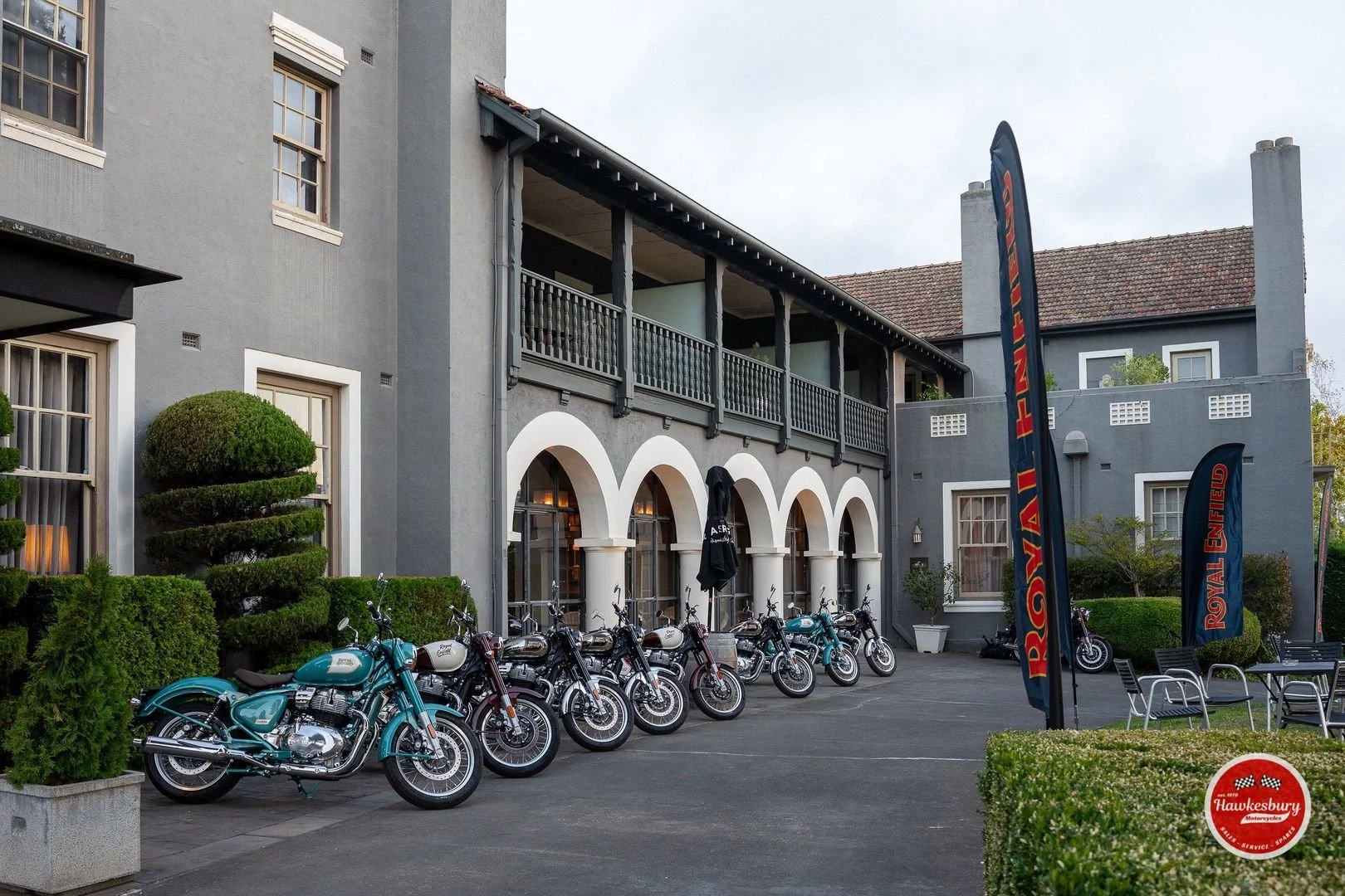 Line of motorcycles parked outside a gray building with arched windows and a balcony. Flags with Royal Enfield branding and Hawkesbury logo are present.