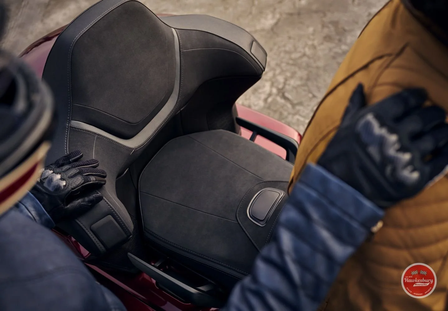 Top-down view of a person wearing black gloves, a brown jacket, and blue jeans, sitting on a red vehicle with a black seat and a matching black storage compartment or tank.