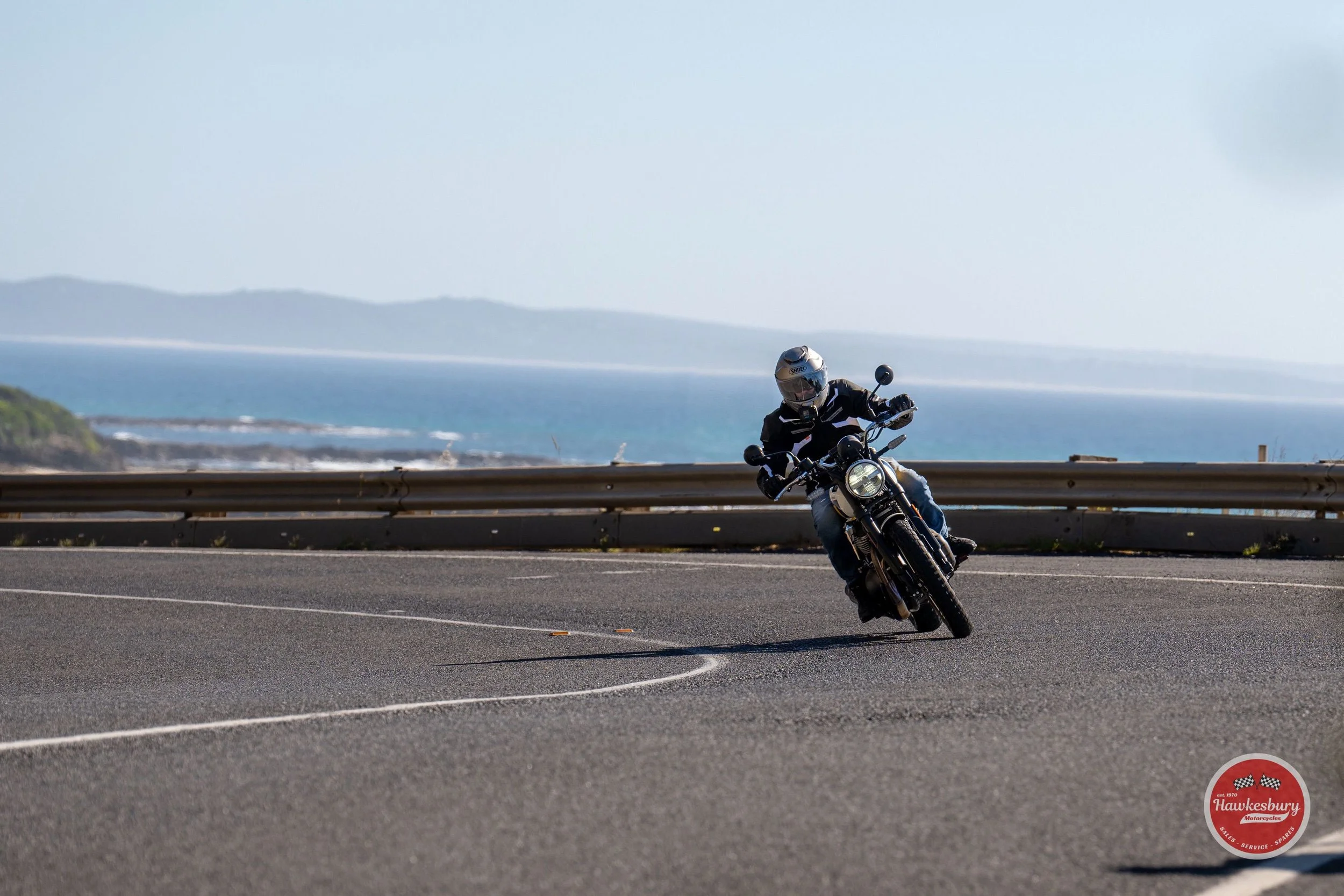 Motorcyclist leaning into a turn on a coastal road with the ocean and distant land in the background.
