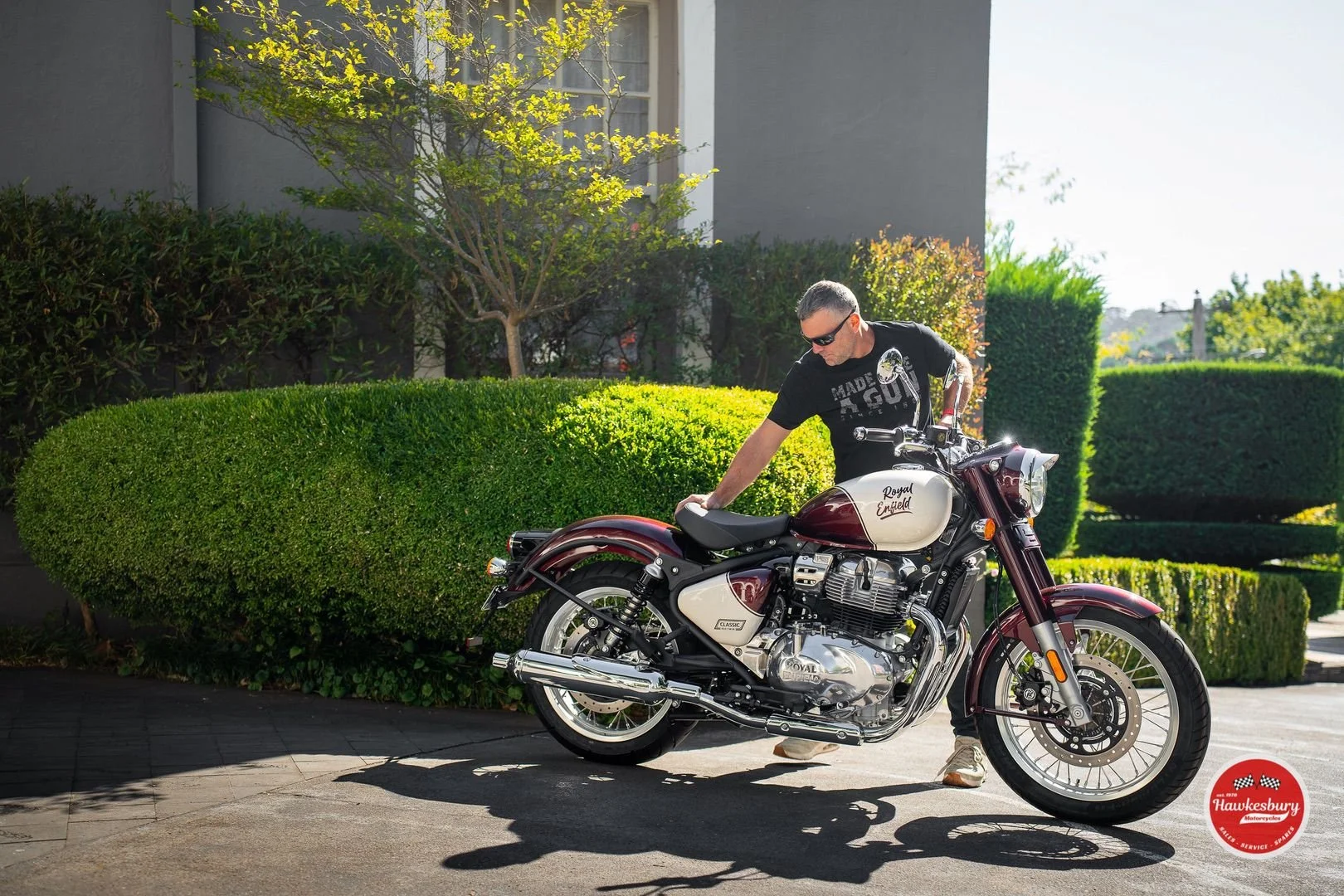 A man in black t-shirt and sunglasses standing beside a classic Royal Enfield motorcycle on a paved driveway, with trimmed green bushes and trees in the background.