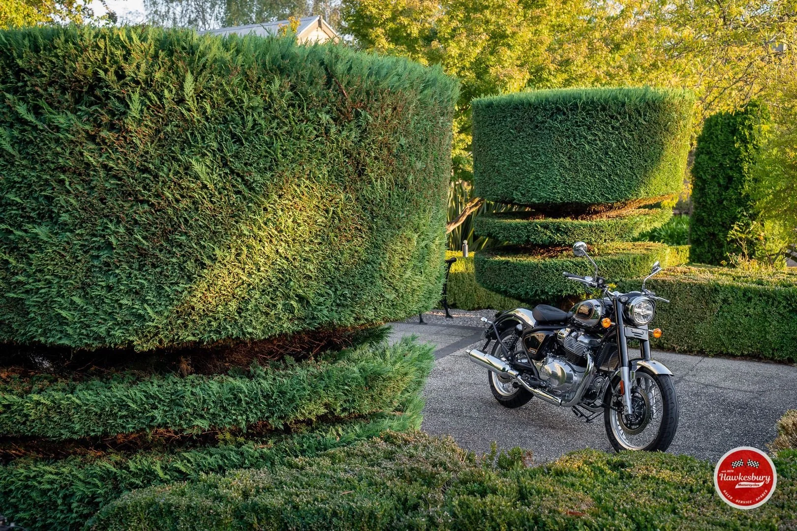 A black motorcycle parked on a paved garden path surrounded by neatly trimmed green hedges and bushes with rounded shapes, under bright sunlight.