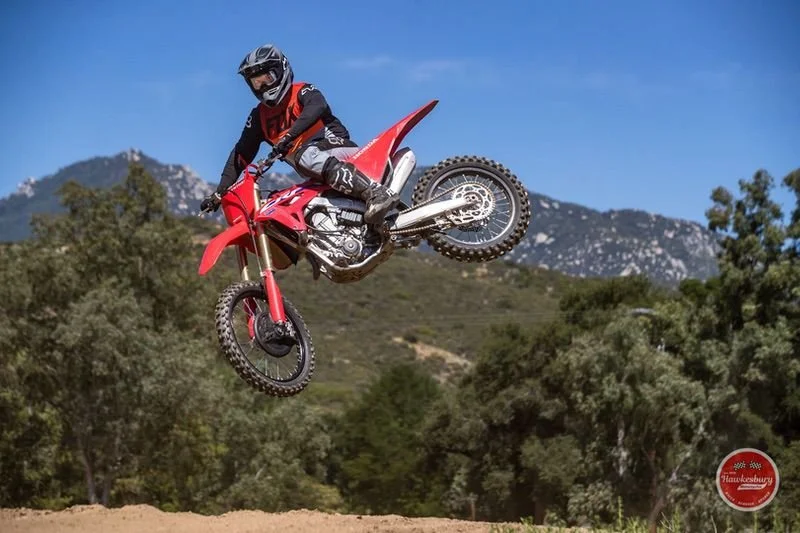 Motocross rider in black and red gear jumps on a red dirt bike over a dirt track with mountains and trees in the background.