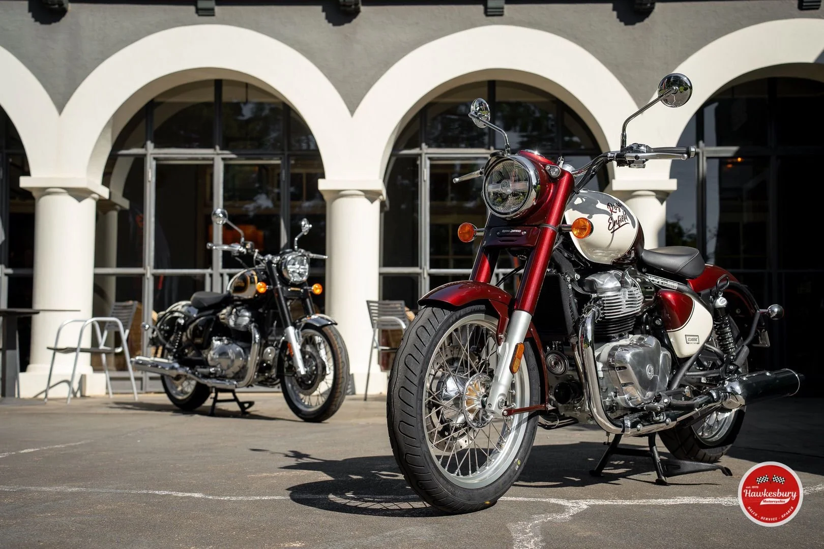 Two vintage motorcycles parked outside a building with arched windows and white columns, with a Hawkesbury logo in the bottom right corner.
