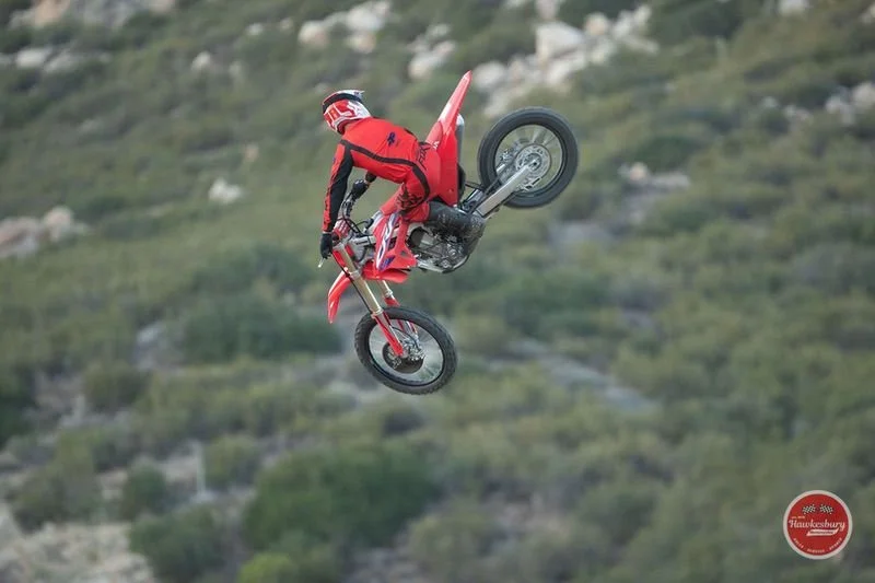 A motocross rider performing a mid-air stunt on a red dirt bike against a backdrop of green hills.