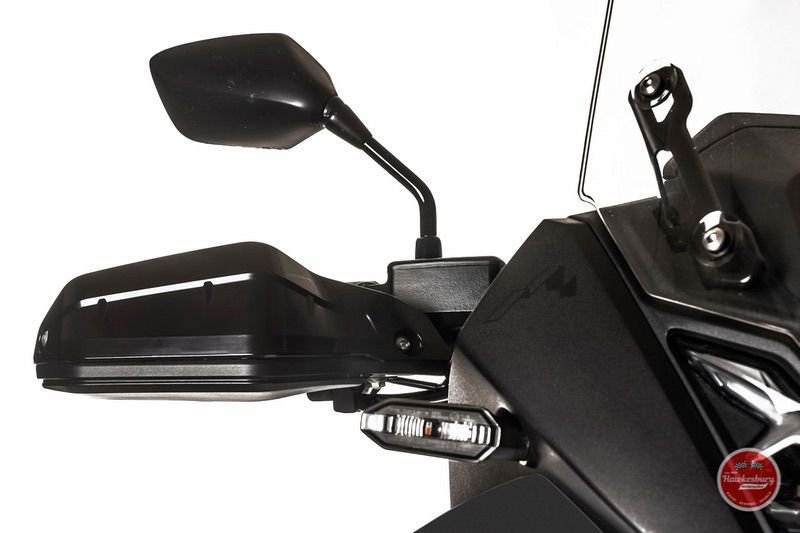 Close-up view of a motorcycle's handlebar, side mirror, and part of the dashboard. The handlebar is black with a side mirror attached, and there's a storage compartment visible. The background is white.