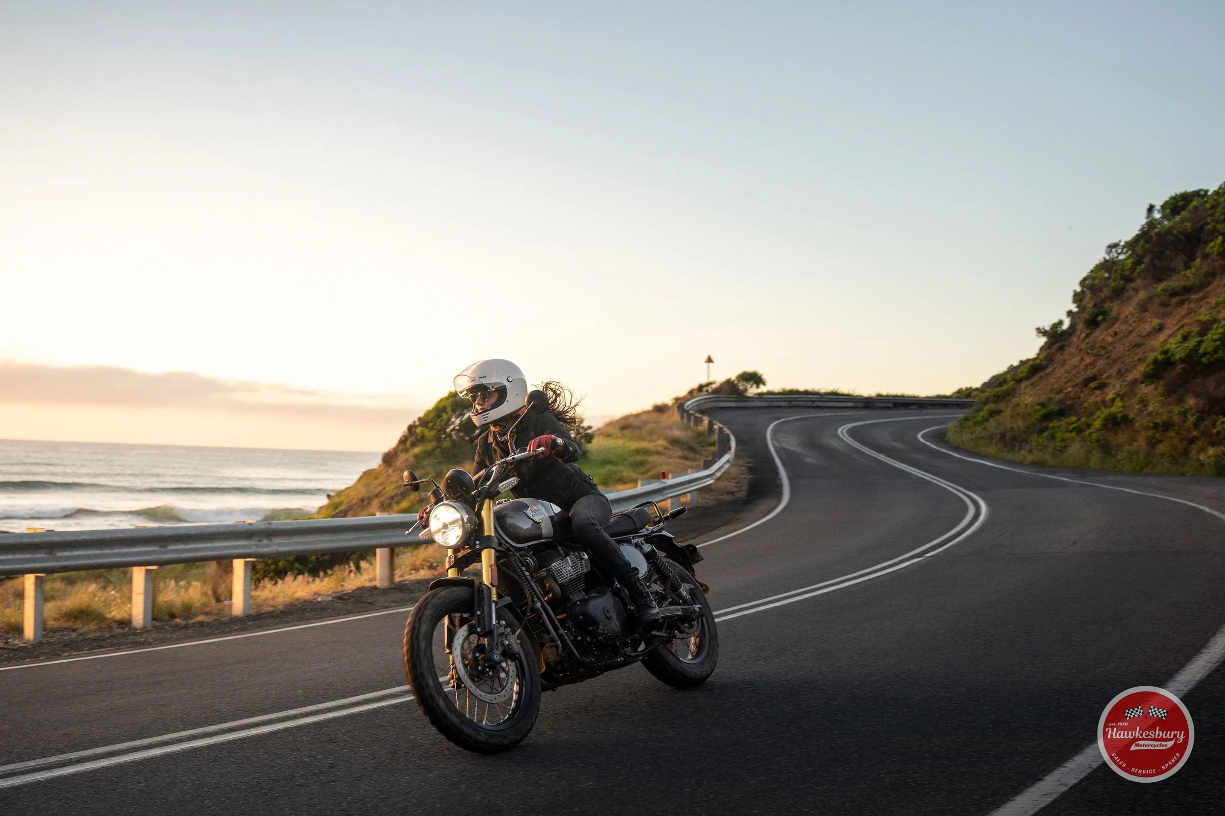 Motorcycle rider wearing a white helmet and black gear riding a black motorcycle on a winding coastal road during sunset, with ocean and hills in the background.
