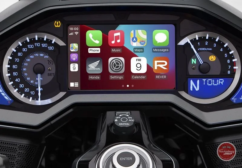 Digital car dashboard display showing app icons for Phone, Music, Maps, Messages, Honda, Settings, Calendar, and REVER, with a speedometer on the left, a fuel gauge and gear indicator on the right, and a central control dial.