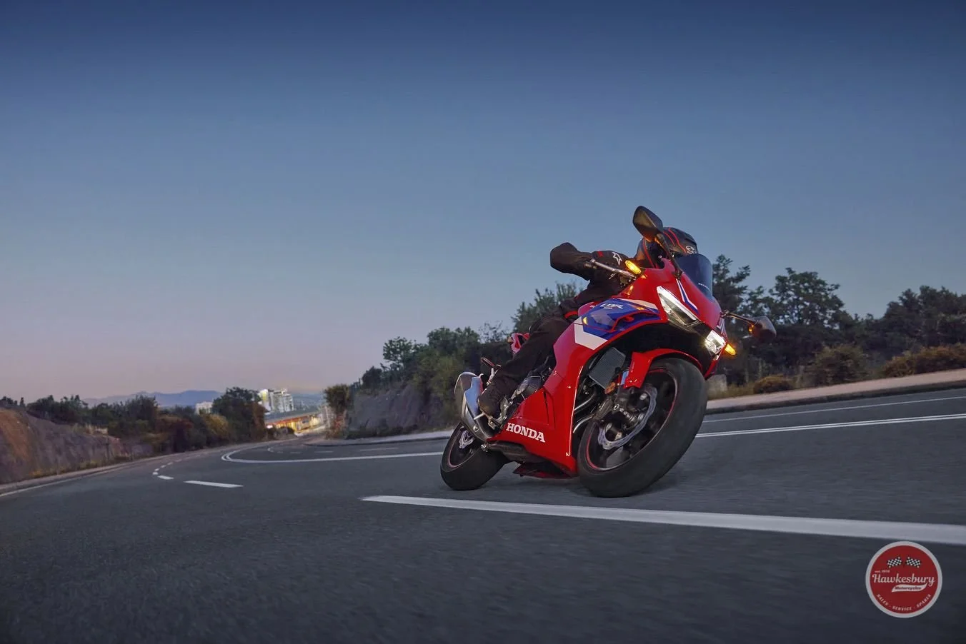 A motorcyclist wearing black gear riding a red and black Honda sport bike on a curving road during dusk with a hills and trees in the background.