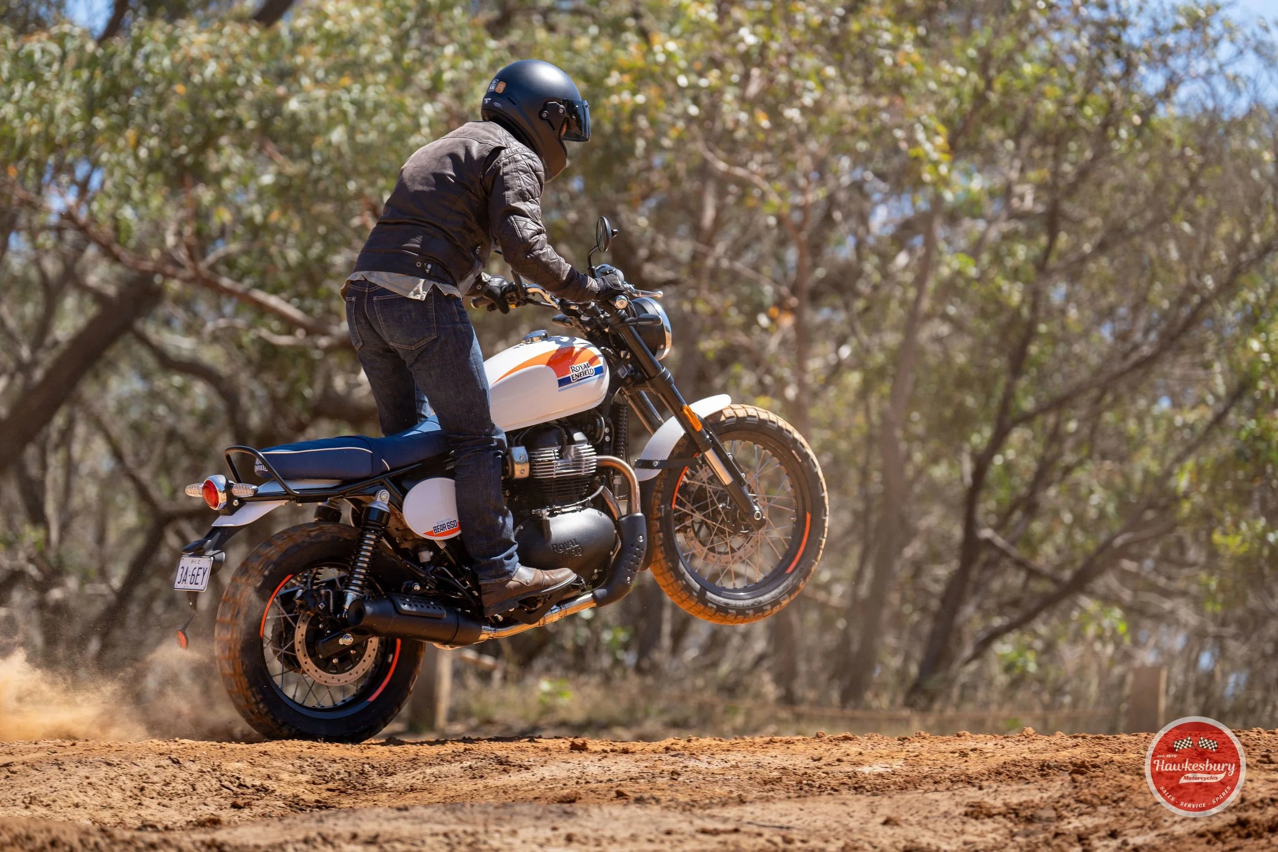 A person riding a vintage-style motorcycle on a dirt path through a wooded area, wearing a helmet, black leather jacket, and jeans.