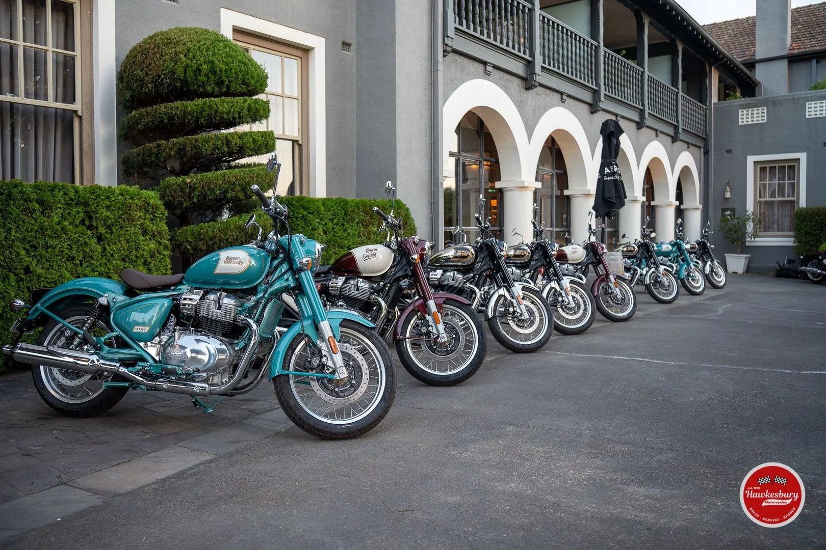 Multiple vintage-style motorcycles parked in a row outside a building with arched windows and a balcony. The motorcycles are mostly turquoise, black, or maroon, with some featuring white accents. There are manicured bushes and a large decorative topi