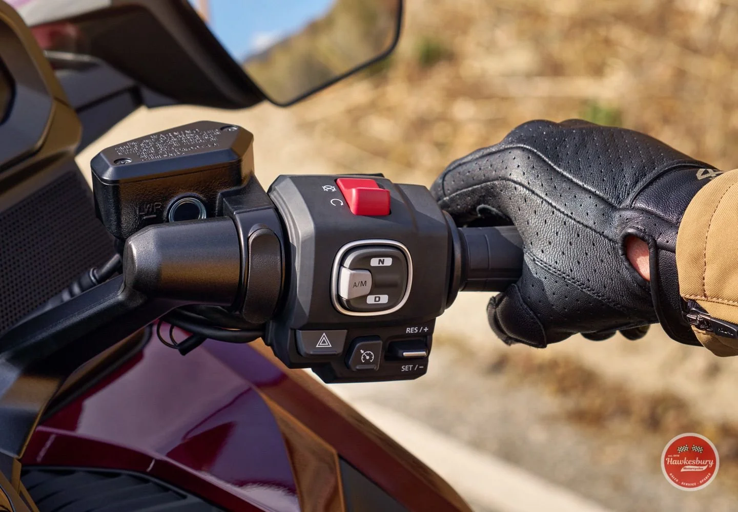 Close-up of a motorcycle handlebar with various control buttons and switches, held by a person wearing a black glove and a beige jacket, with a blurred outdoor background.