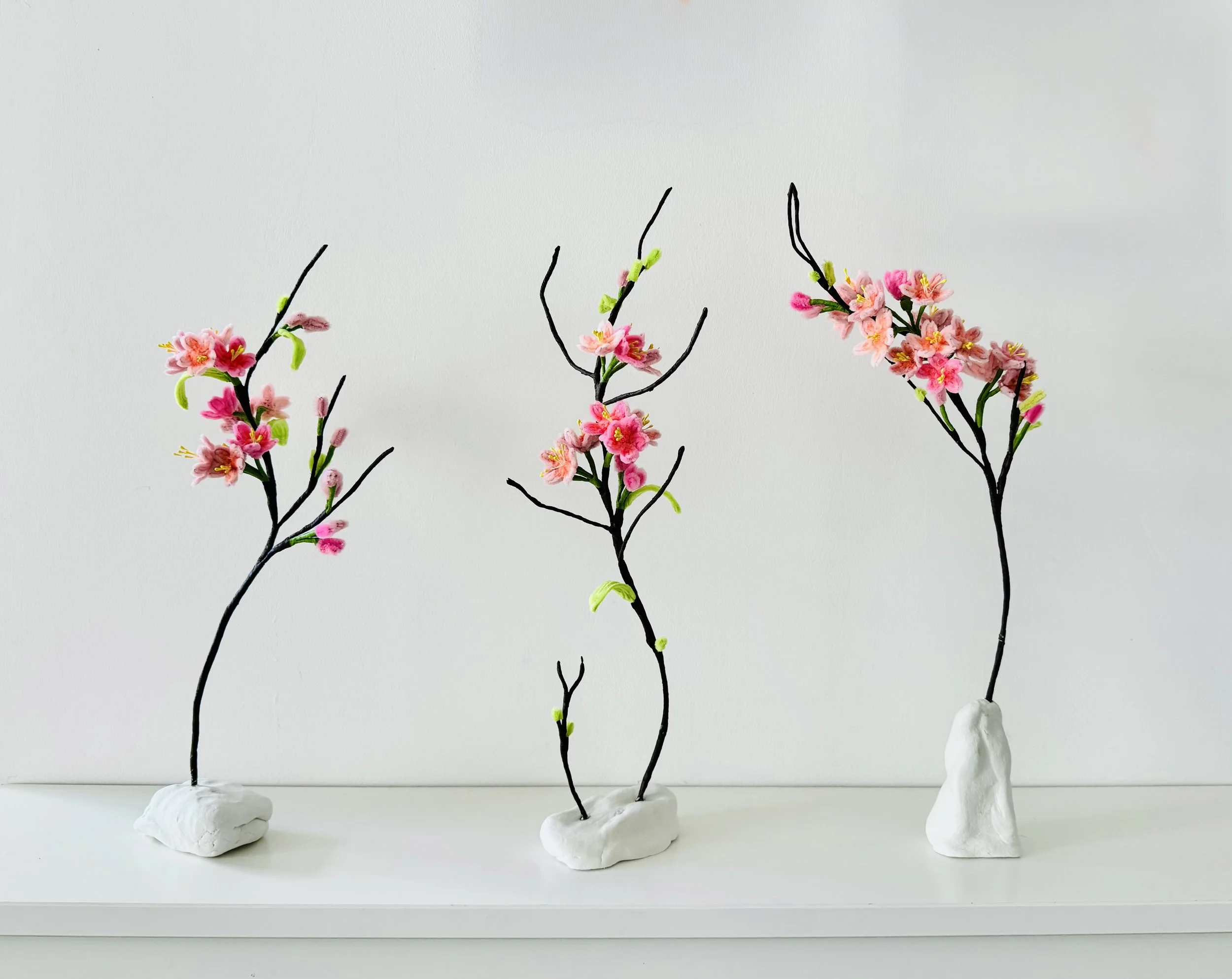Three minimalist flower arrangements with pink blossoms on black wire branches, each secured in white clay bases, placed on a white surface against a plain white background.
