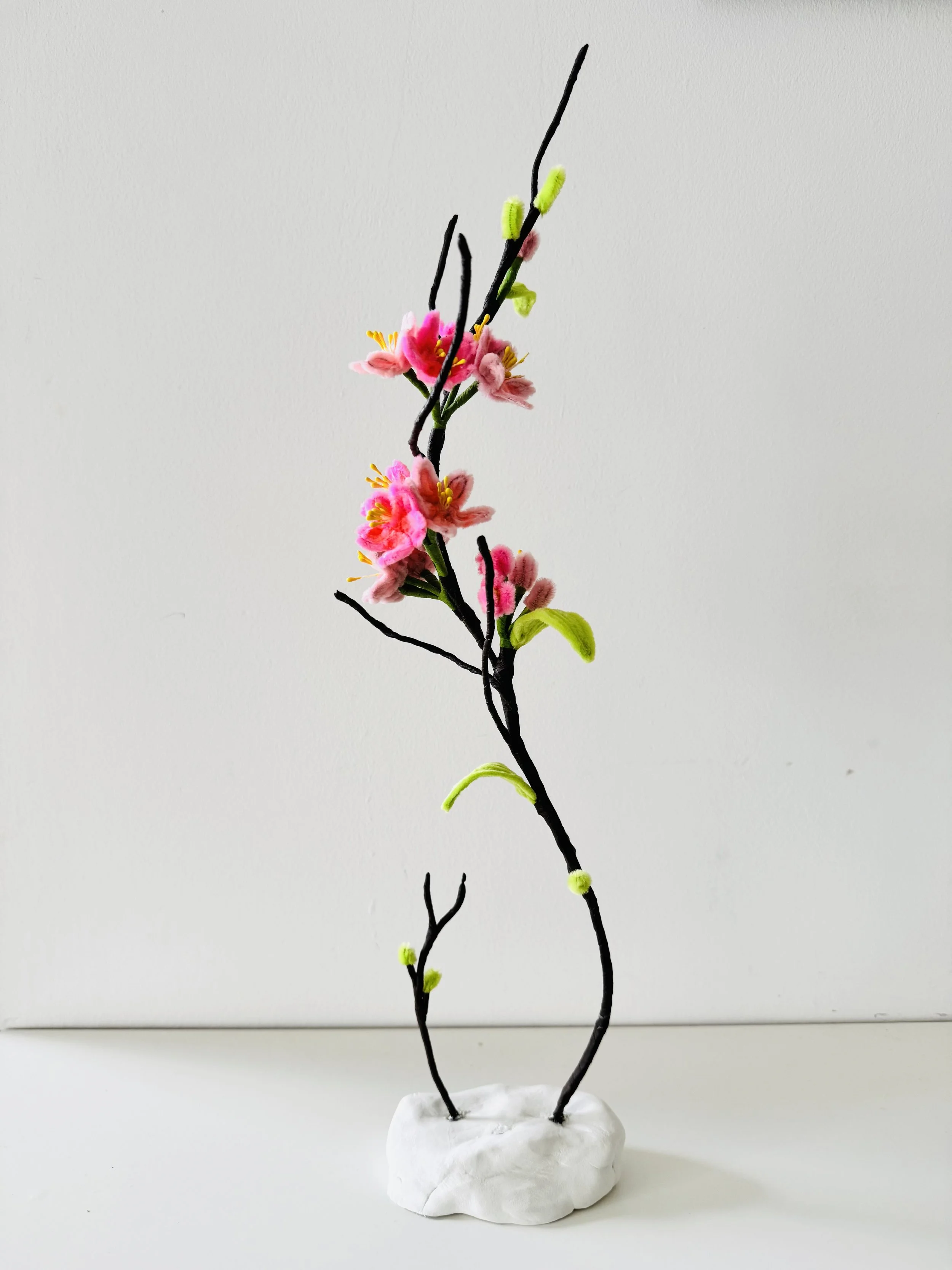 A handmade decorative piece resembling a flowering branch with pink blossoms, green leaves, and black twigs, mounted on a white base.