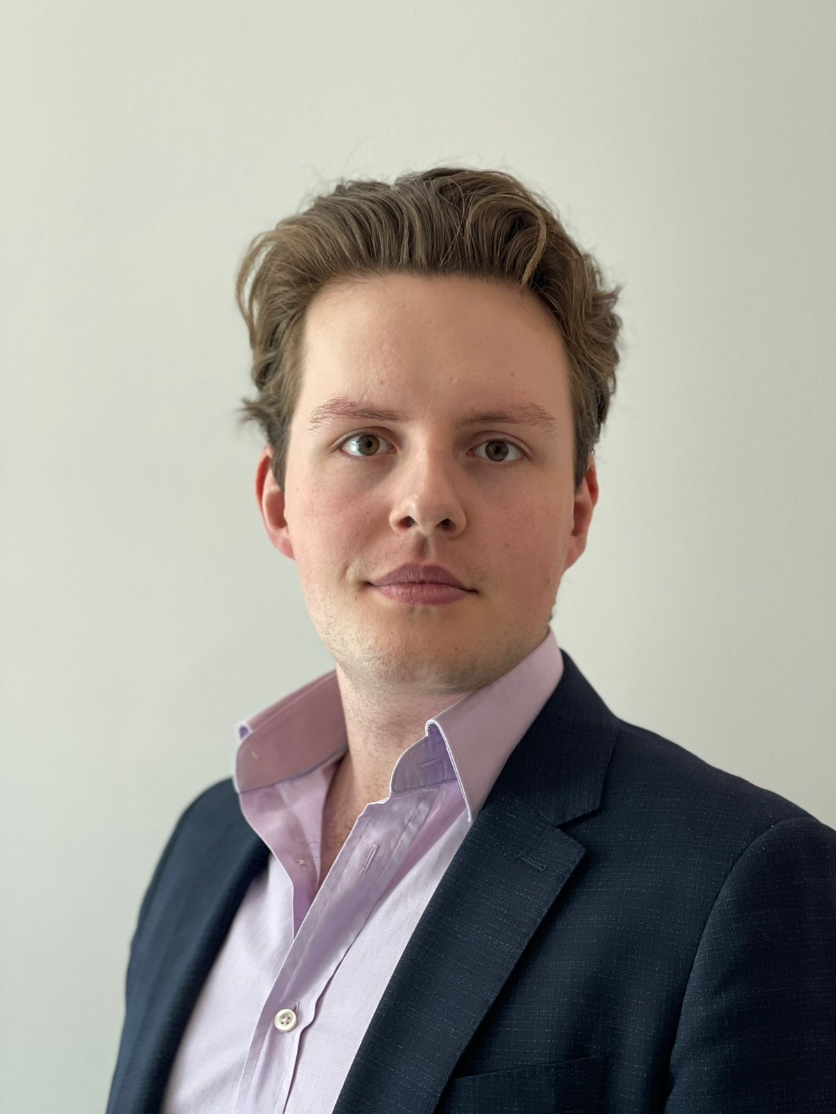 A young man with wavy brown hair, wearing a dark suit jacket and a light pink dress shirt, looking directly at the camera with a neutral expression against a plain white background.