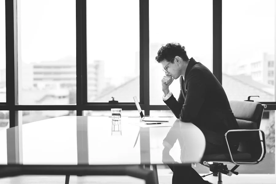A man in a suit sitting at a desk in an office, looking at a laptop, with a glass of water in front of him, in front of large windows showing a cityscape.