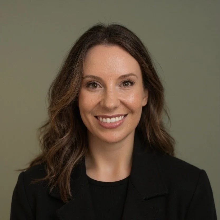 A woman with wavy brown hair wearing a black blazer and smiling against a plain green background.
