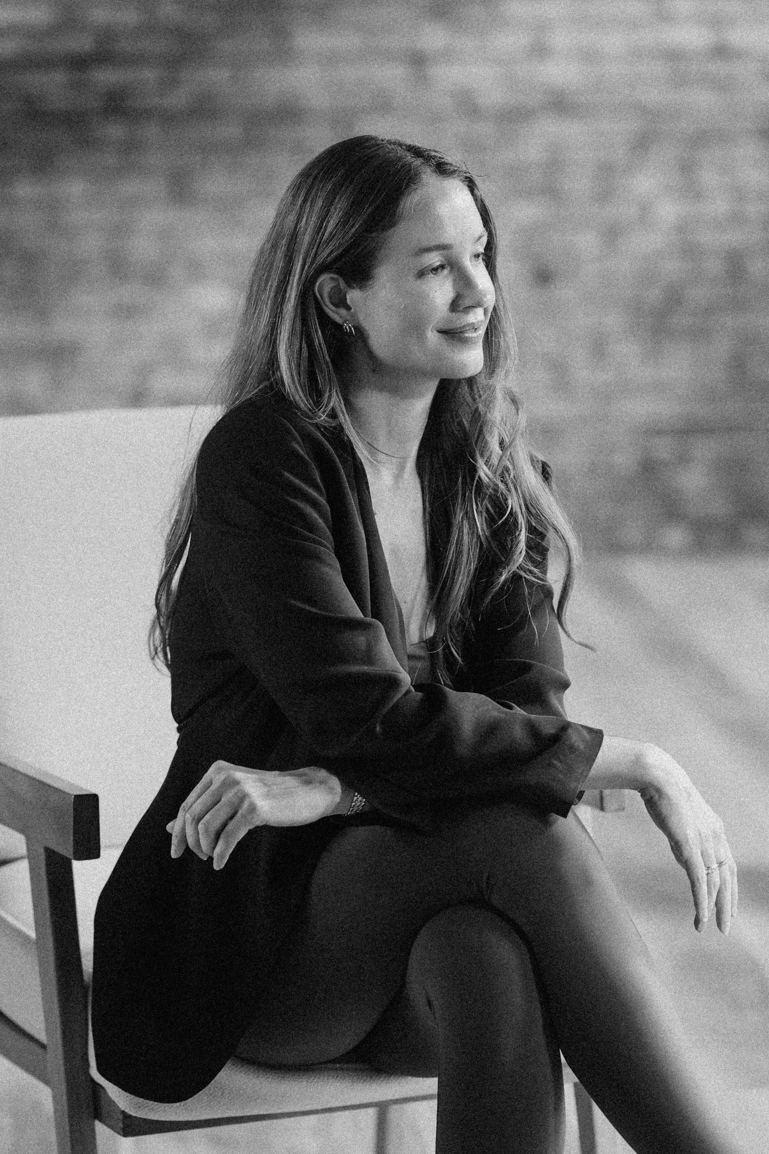 Black-and-white photo of a woman sitting on a chair, wearing a blazer and pants, with long wavy hair, smiling slightly, looking to the side.
