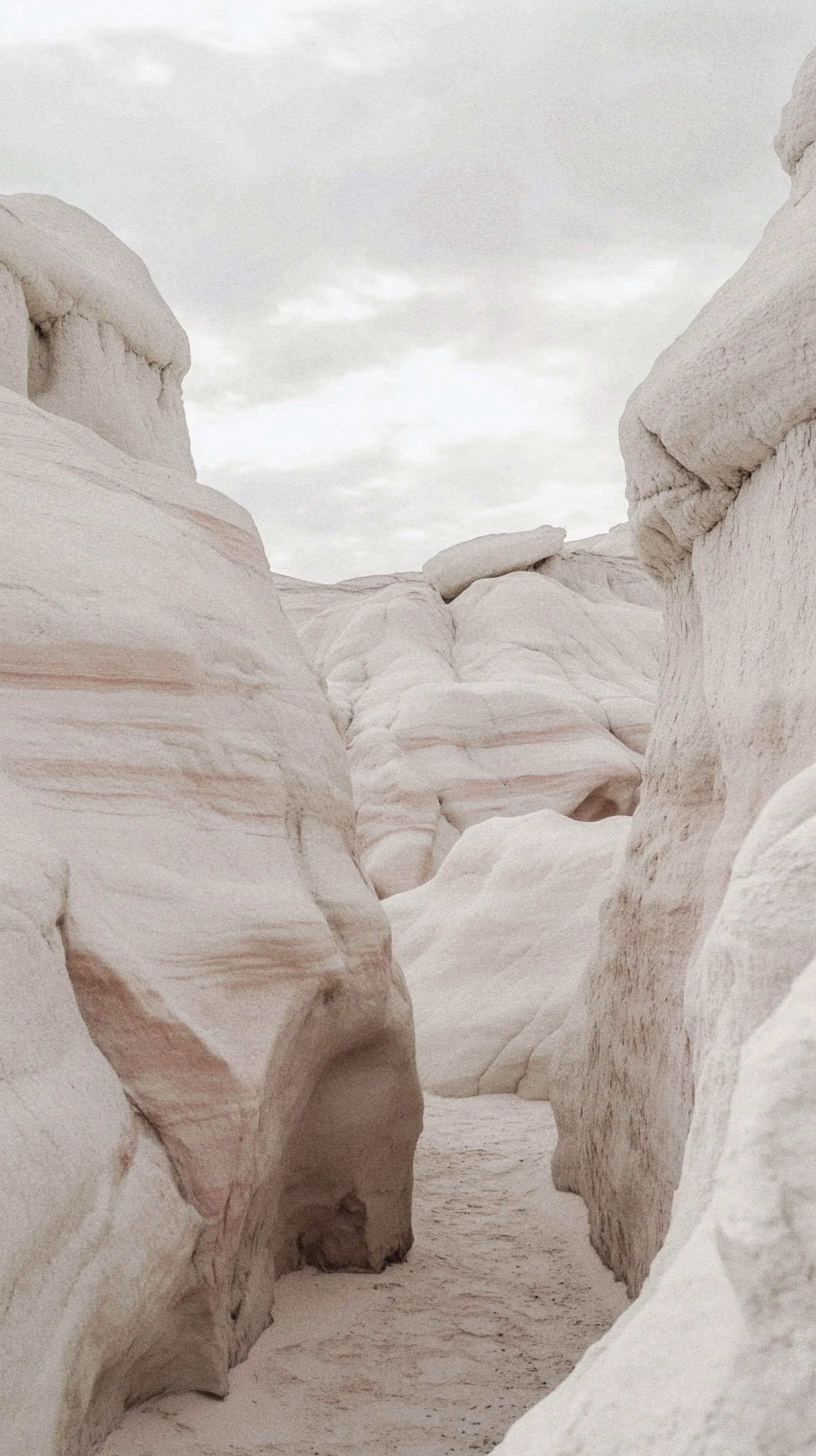 Narrow canyon with smooth, layered white and pinkish rock formations under a cloudy sky.