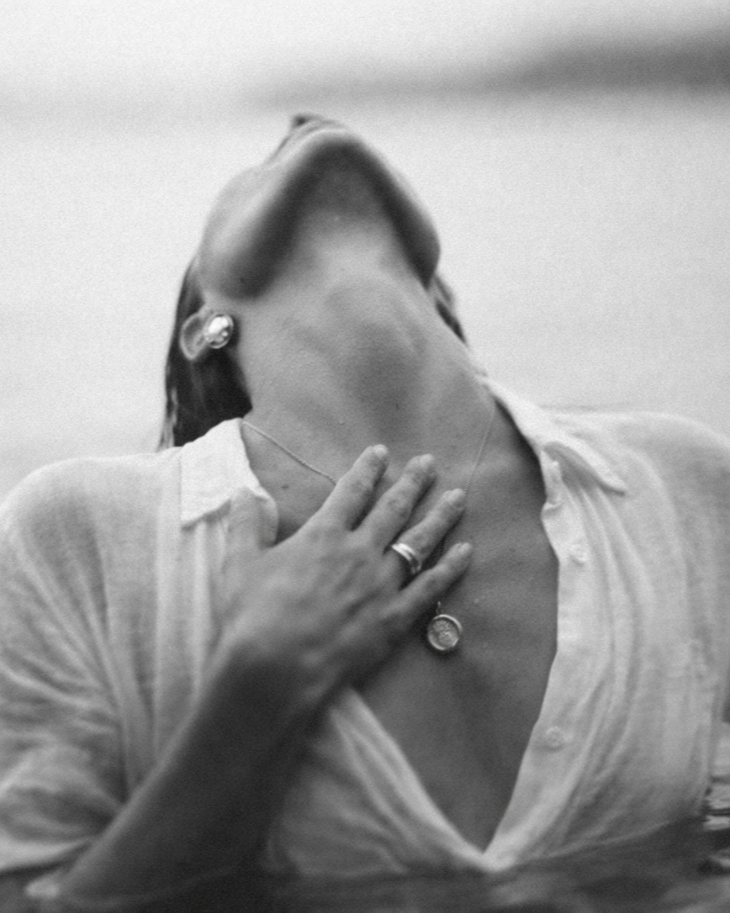 Black and white photo of a woman tilting her head back with eyes closed, touching her neck with her left hand, wearing a white blouse and jewelry.
