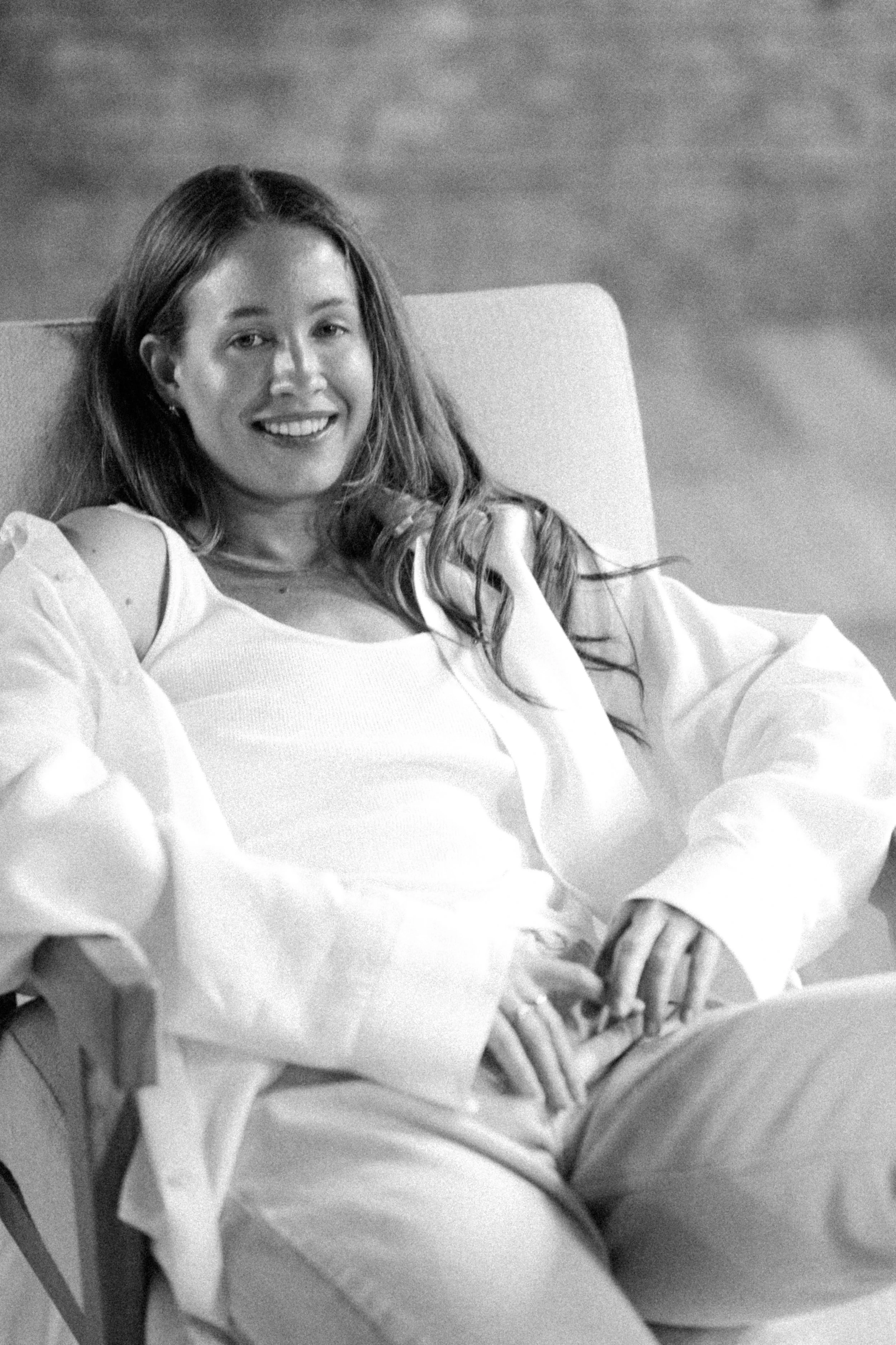 A smiling woman with long hair, wearing a light-colored top, sitting on a chair with her hands resting on her lap.