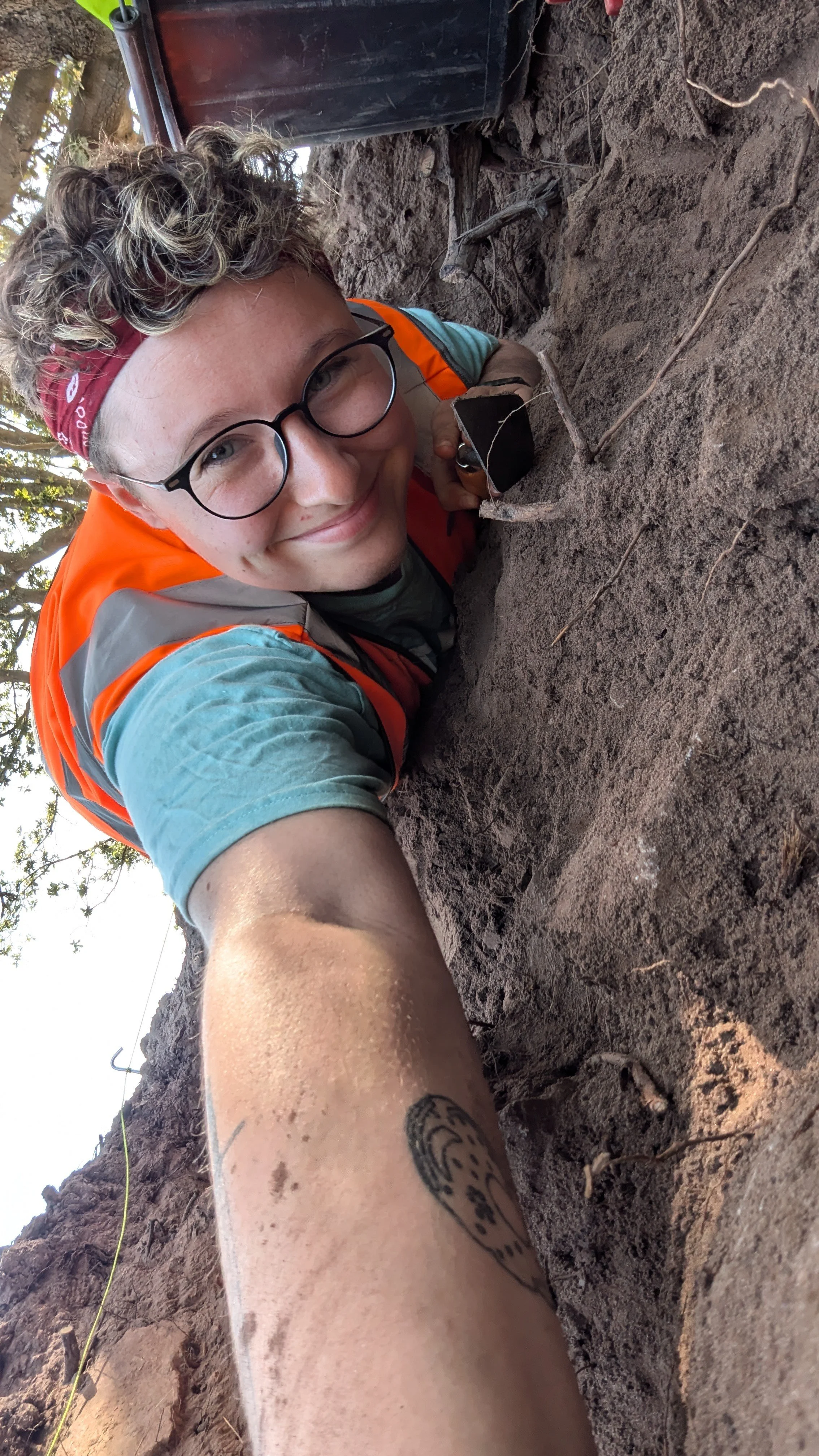 A smiling Louise Bedford (@louisearchaeology) with curly hair, glasses, and a red bandana, lying on dirt ground with a camera near her face. She is wearing an orange safety vest and a teal t-shirt, and is excavating a trench.