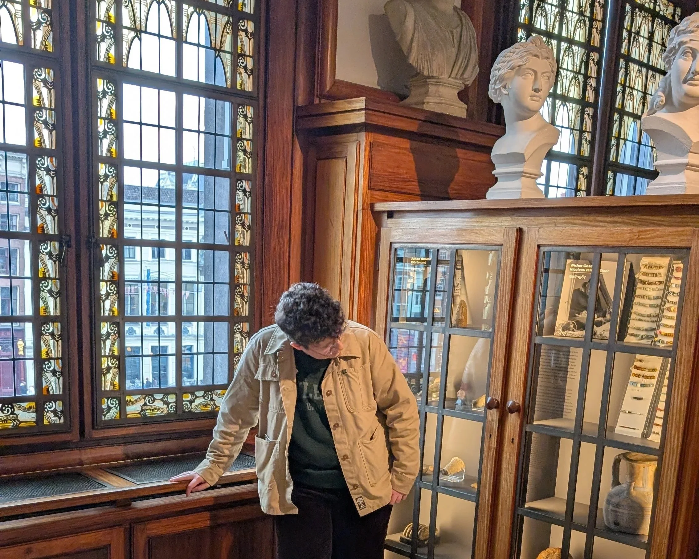 Louise Bedford (@louisearchaeology) is central. She has short, brown curly hair and wears glasses. She is looking down, wearing a beige jacket in a museum with large glass windows, wooden display cases, and classical bust sculptures.