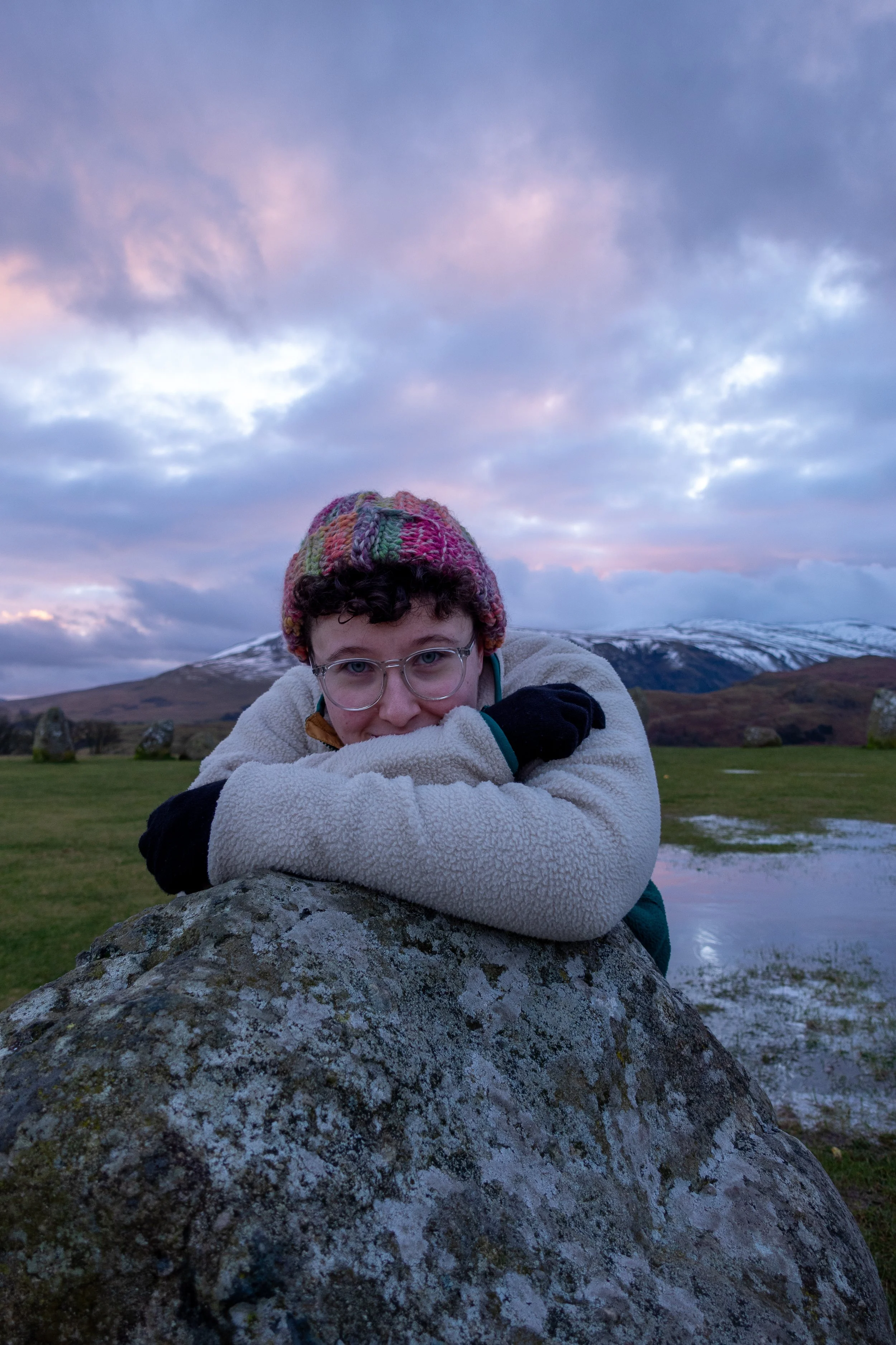 Louise Bedford (@louisearchaeology) is central. She has short, brown curly hair and wears glasses. She wears a colourful knit hat and warm clothing, leaning on a standing stone, with Castlerigg stone circle in the background